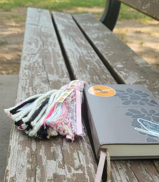 bible with bible tassel on a park bench. Featuring rainbow, and pink ribbon/ yarn